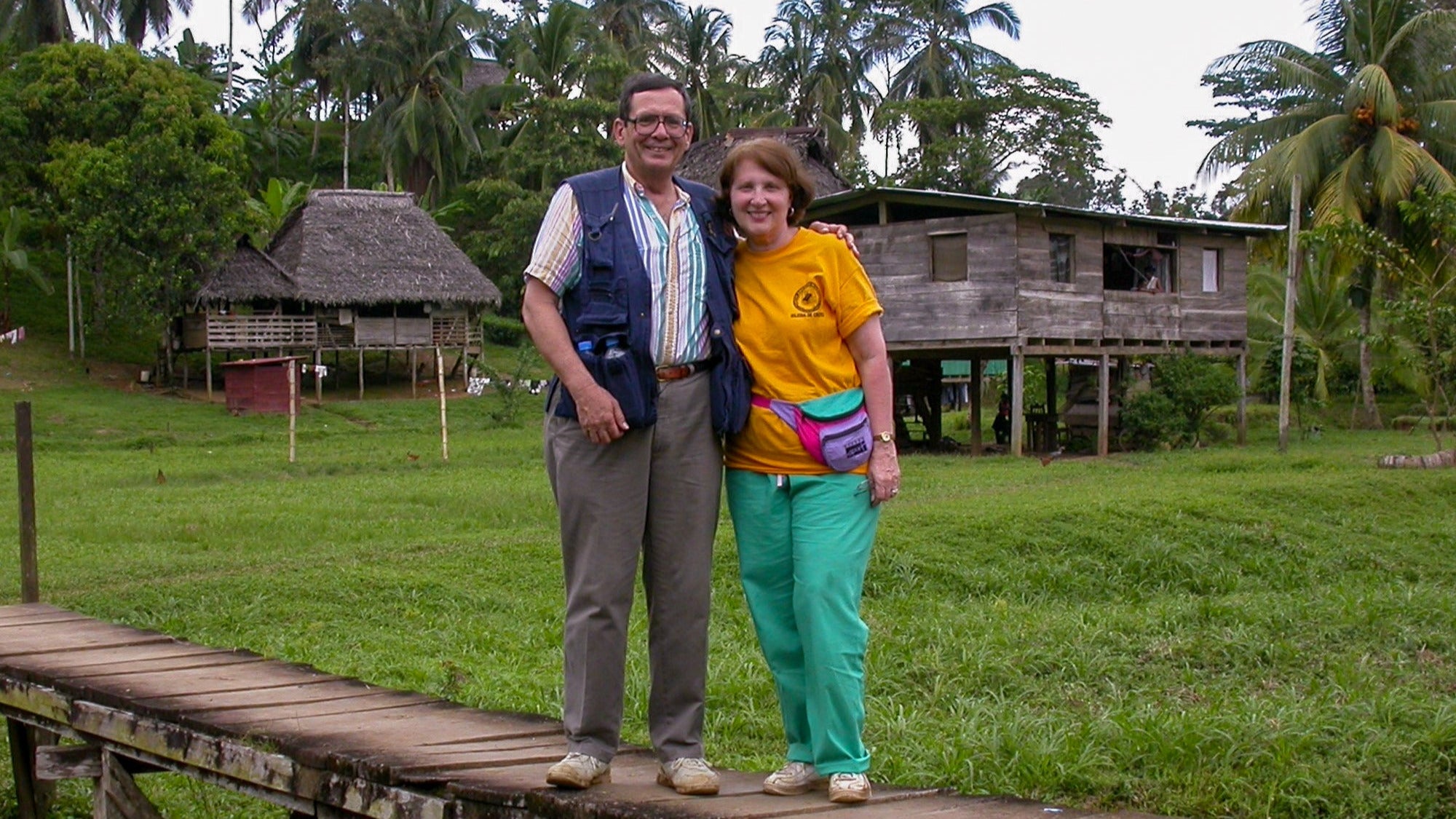 Two people standing on a wooden walkway with tropical huts and greenery in the background