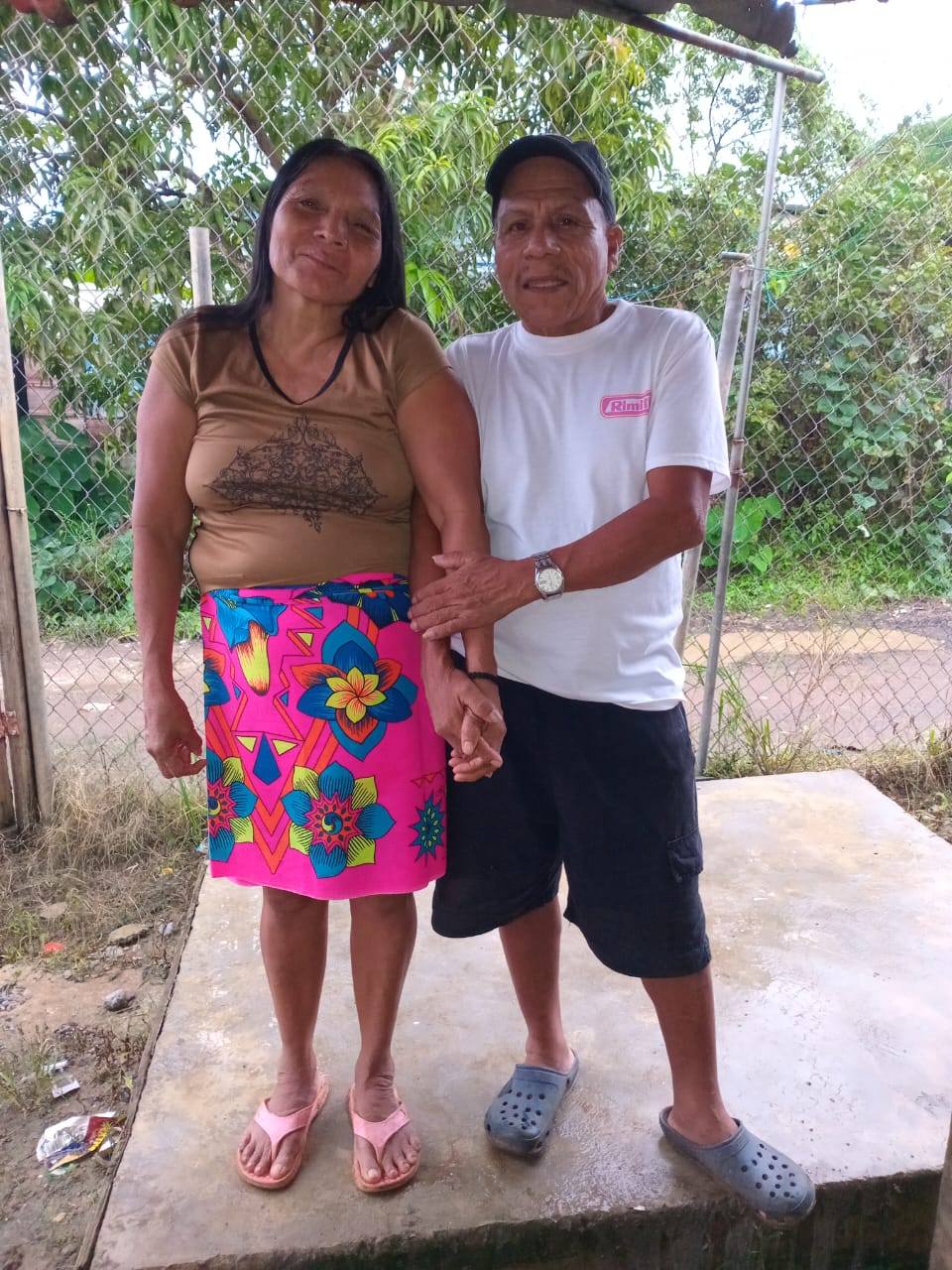 Two people standing together outdoors with a chain-link fence and greenery in the background.